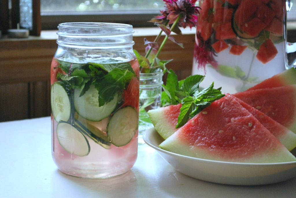 Bee balm, cucumber, and watermelon infused water in a mason jar with watermelon slices and a pitcher of water nearby.